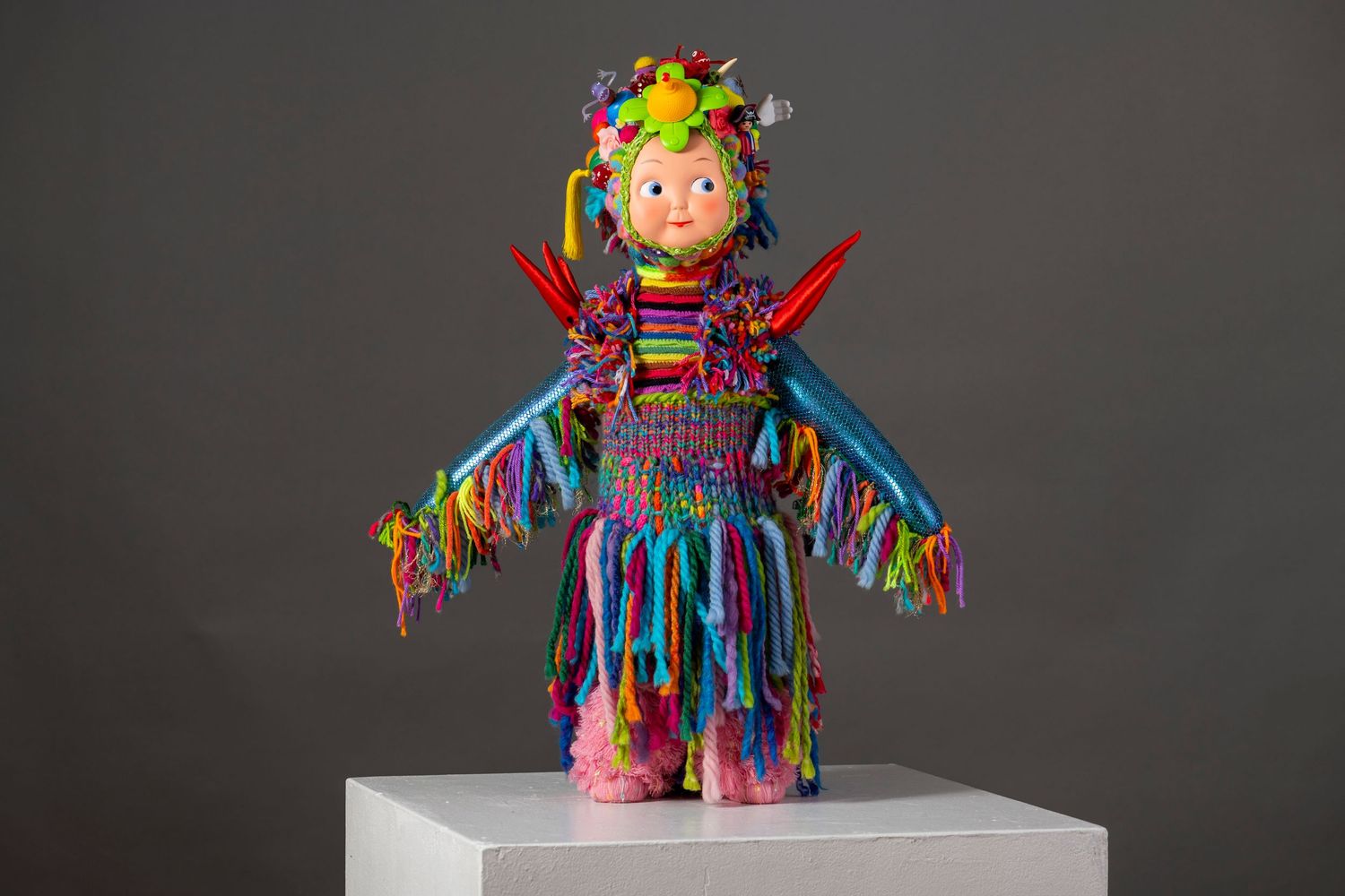 Colorful plush toy doll wearing vibrant rainbow fringe costume and flower crown stands on white pedestal against gray background.