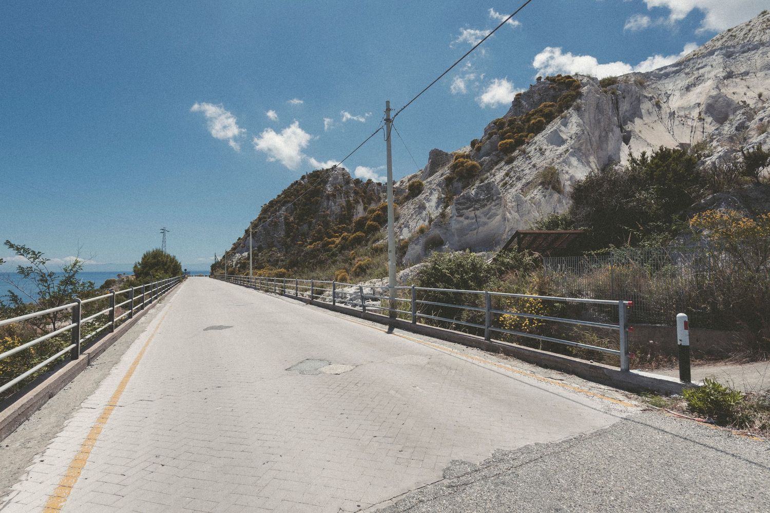 A winding mountain road with metal guardrails curves along steep rocky cliffs against a bright blue sky.