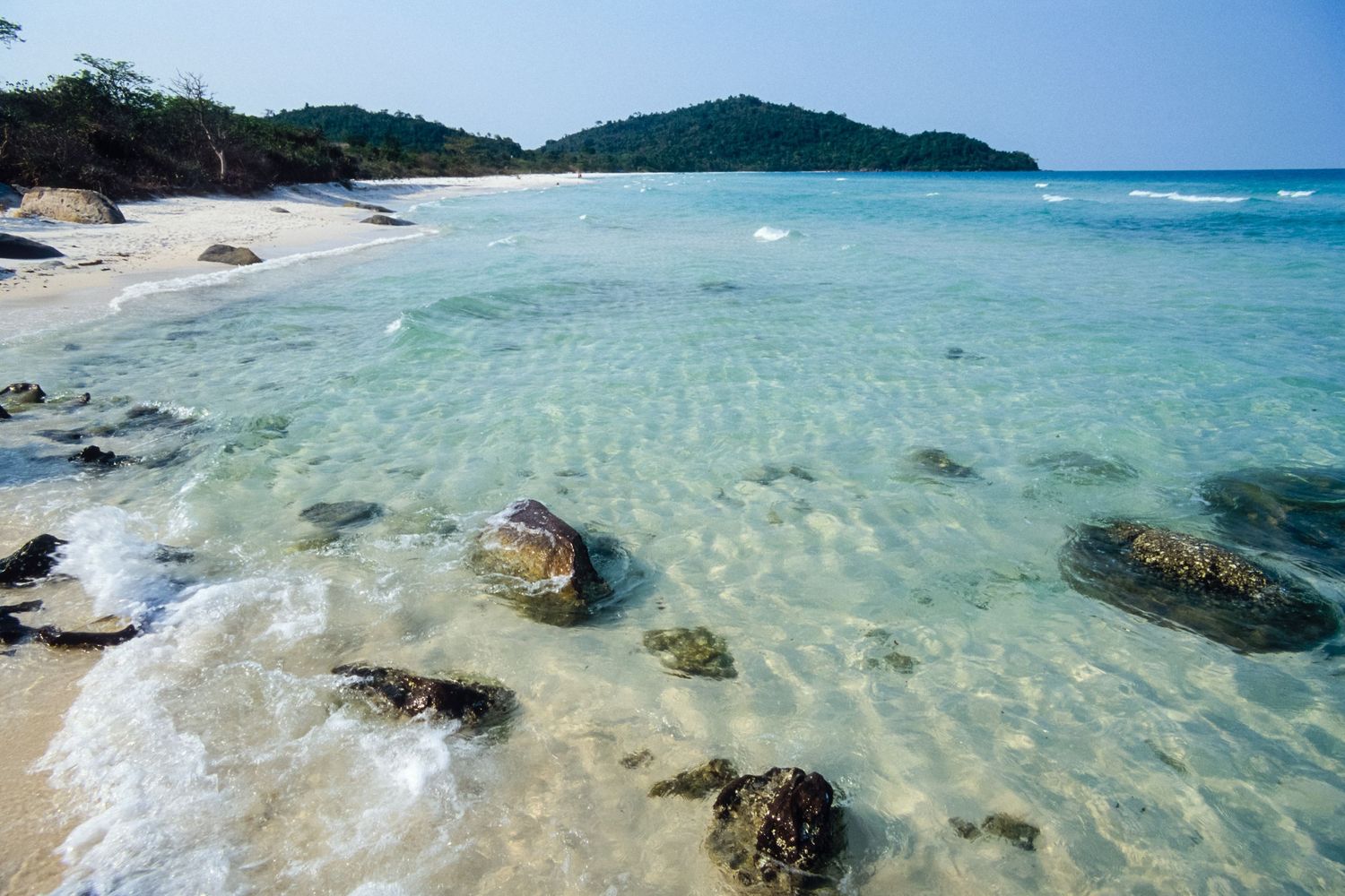 Crystal clear turquoise waters meet white sandy beaches along a tropical coastline with rocky outcrops.