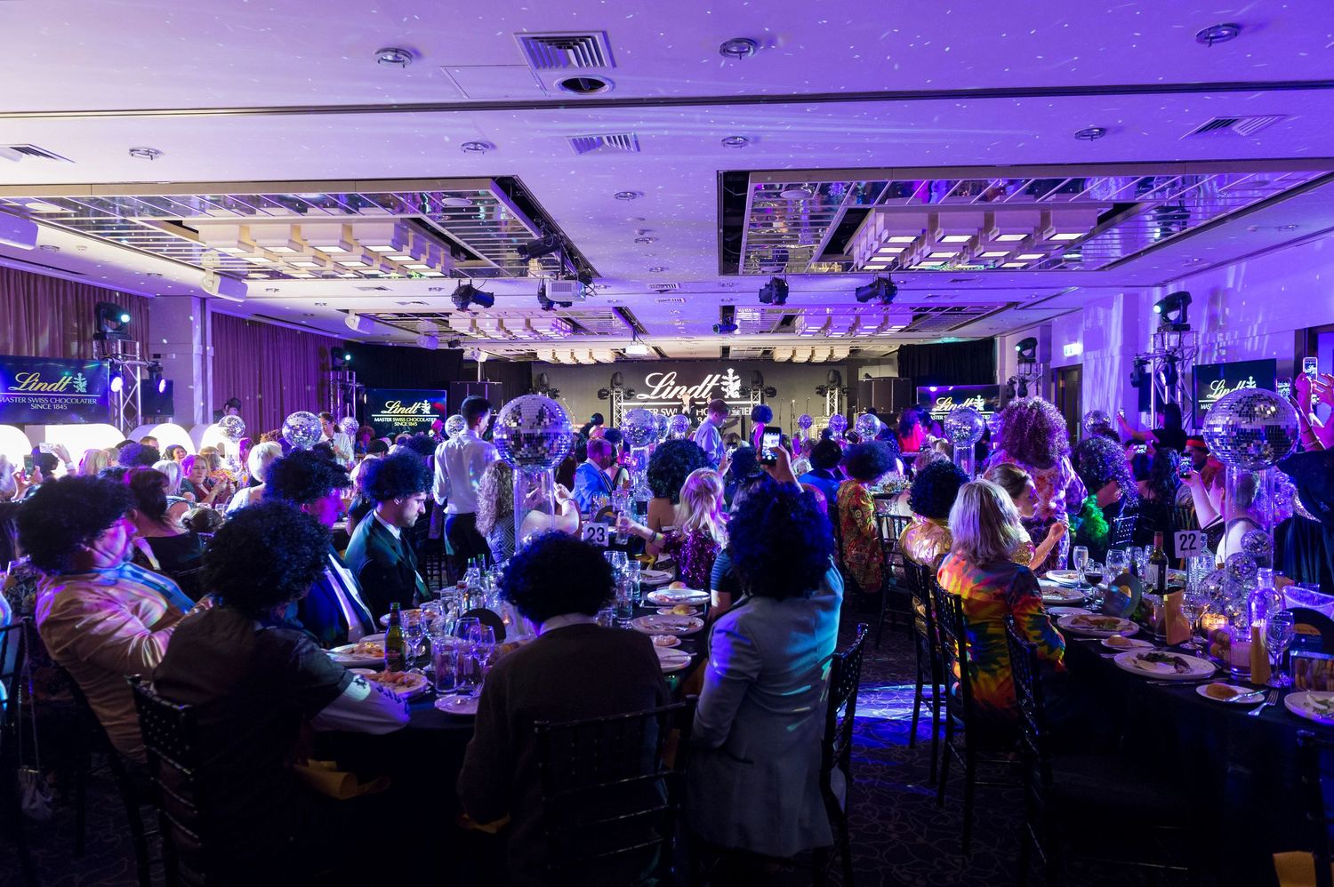 Large crowd of people gathered in a dimly lit banquet hall with purple lighting and tables during an evening event.