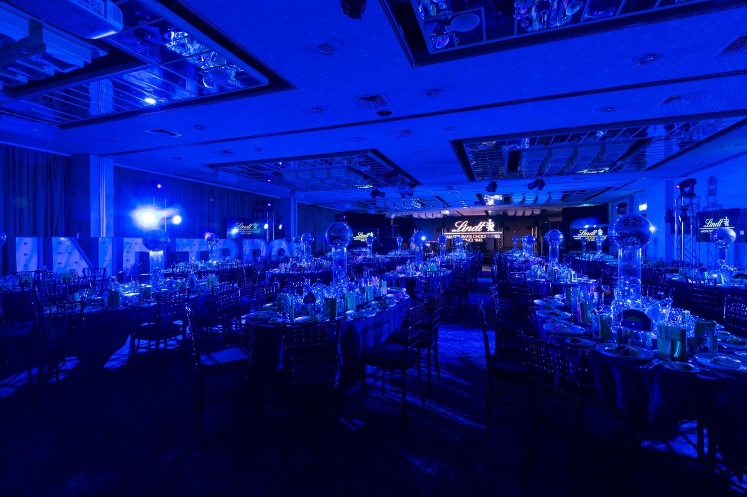 A large ballroom illuminated in deep blue lighting with elegant table settings and dramatic ceiling features.