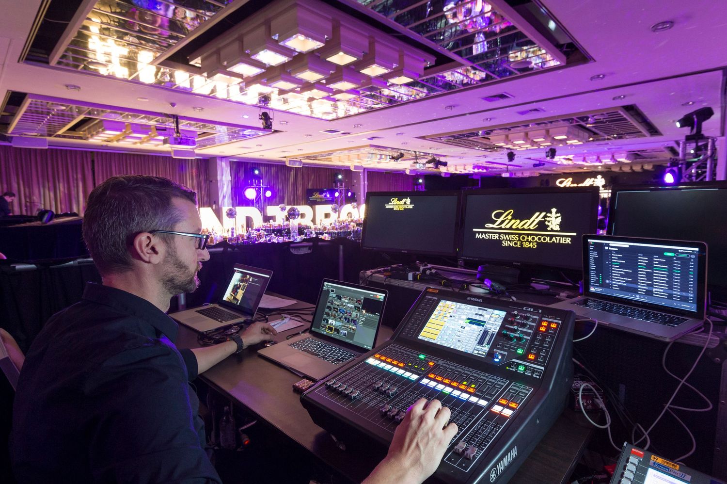 Professional DJ equipment setup in a dark room illuminated by purple lighting showing mixing boards and controllers.