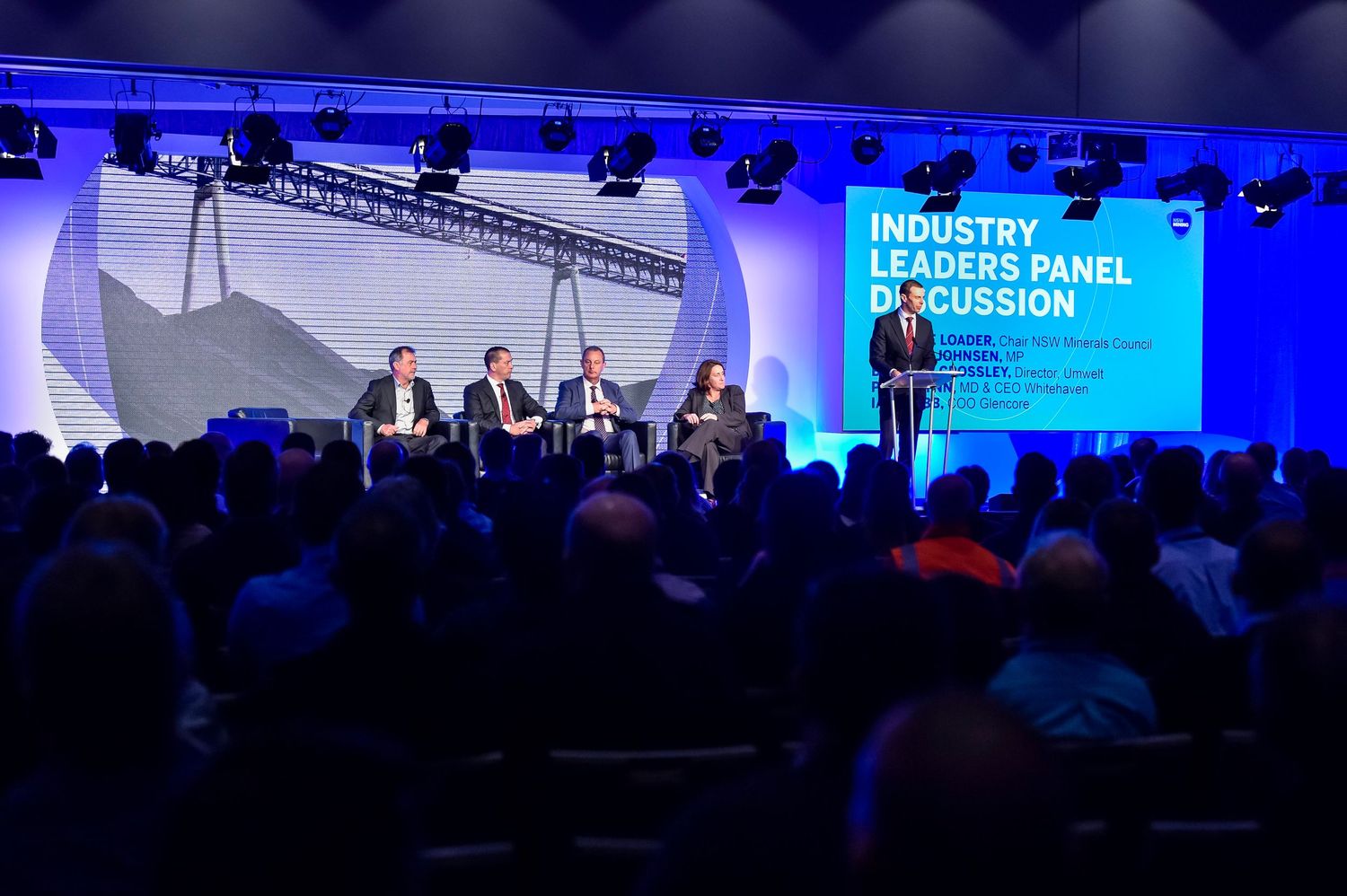 Business conference stage with panel discussion setup and blue ambient lighting.