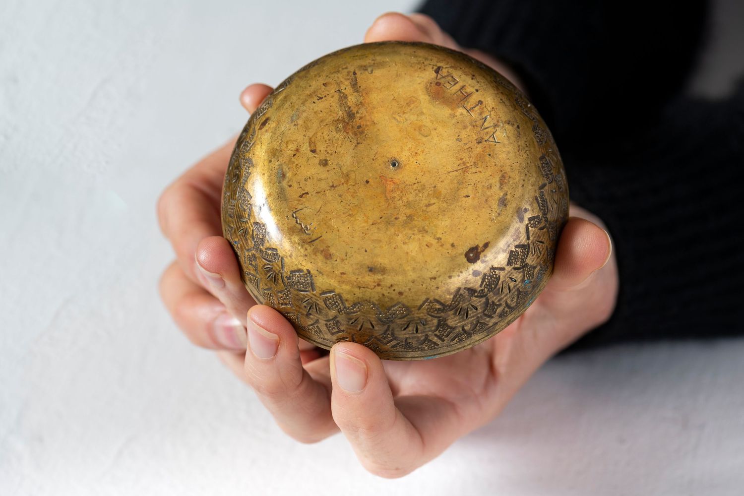 Hands holding and rotating a weathered gold-colored spherical object against a dark background.