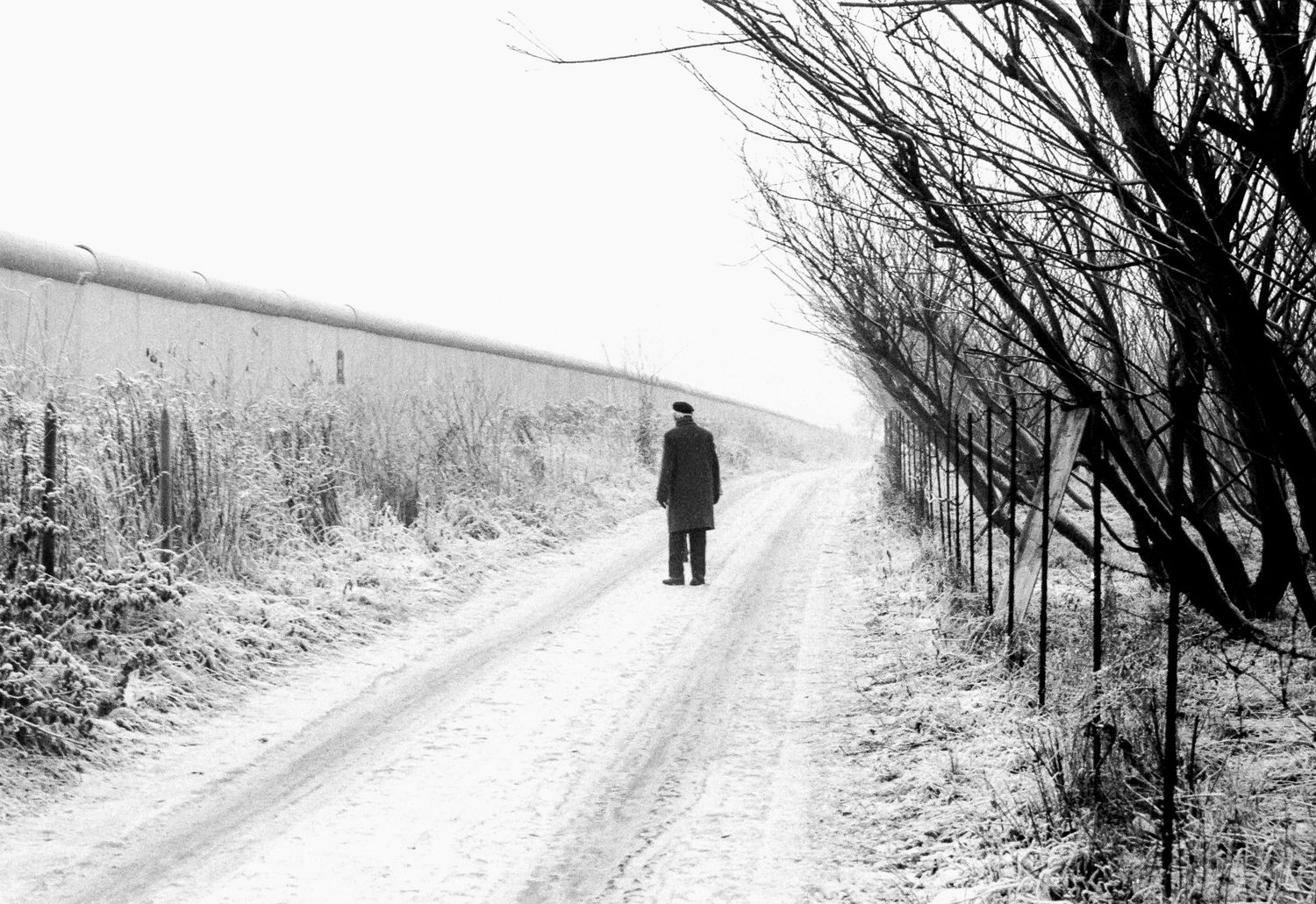 Snow-covered path along the Berlin Wall with a lone figure in the distance.