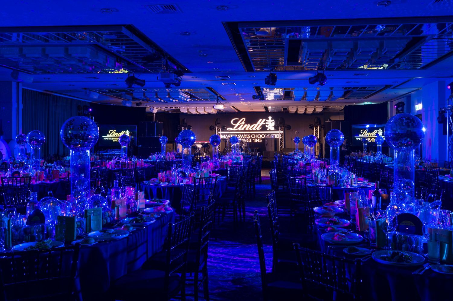 A large banquet hall illuminated in vibrant blue lighting with tables and chairs arranged for an elegant event.