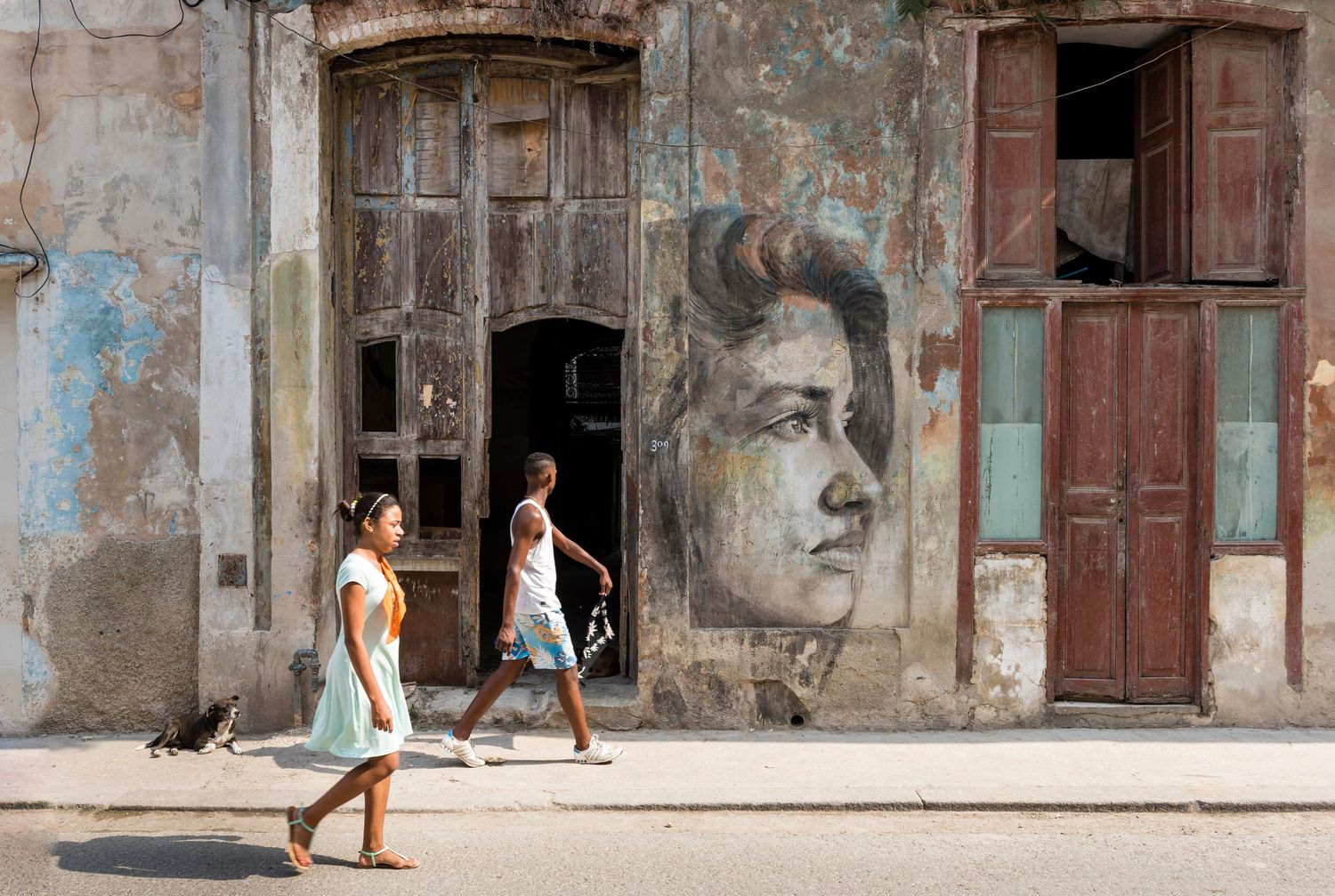 Street scene with large portrait mural painted on weathered building wall.