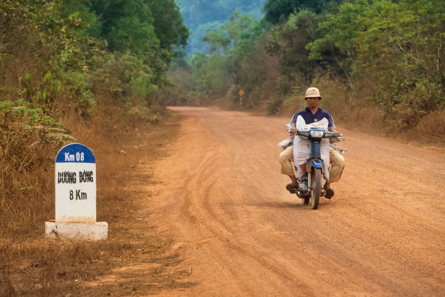 Dusty dirt road winding through rural countryside with motorcycles in distance.