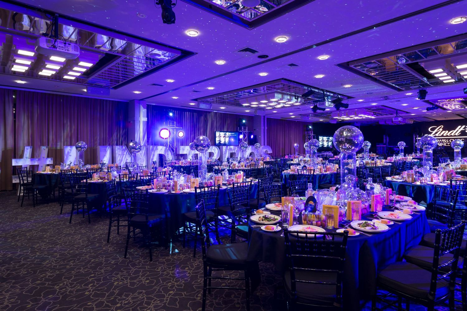 A large banquet hall illuminated in dramatic purple and blue lighting with elegant table settings and decor.