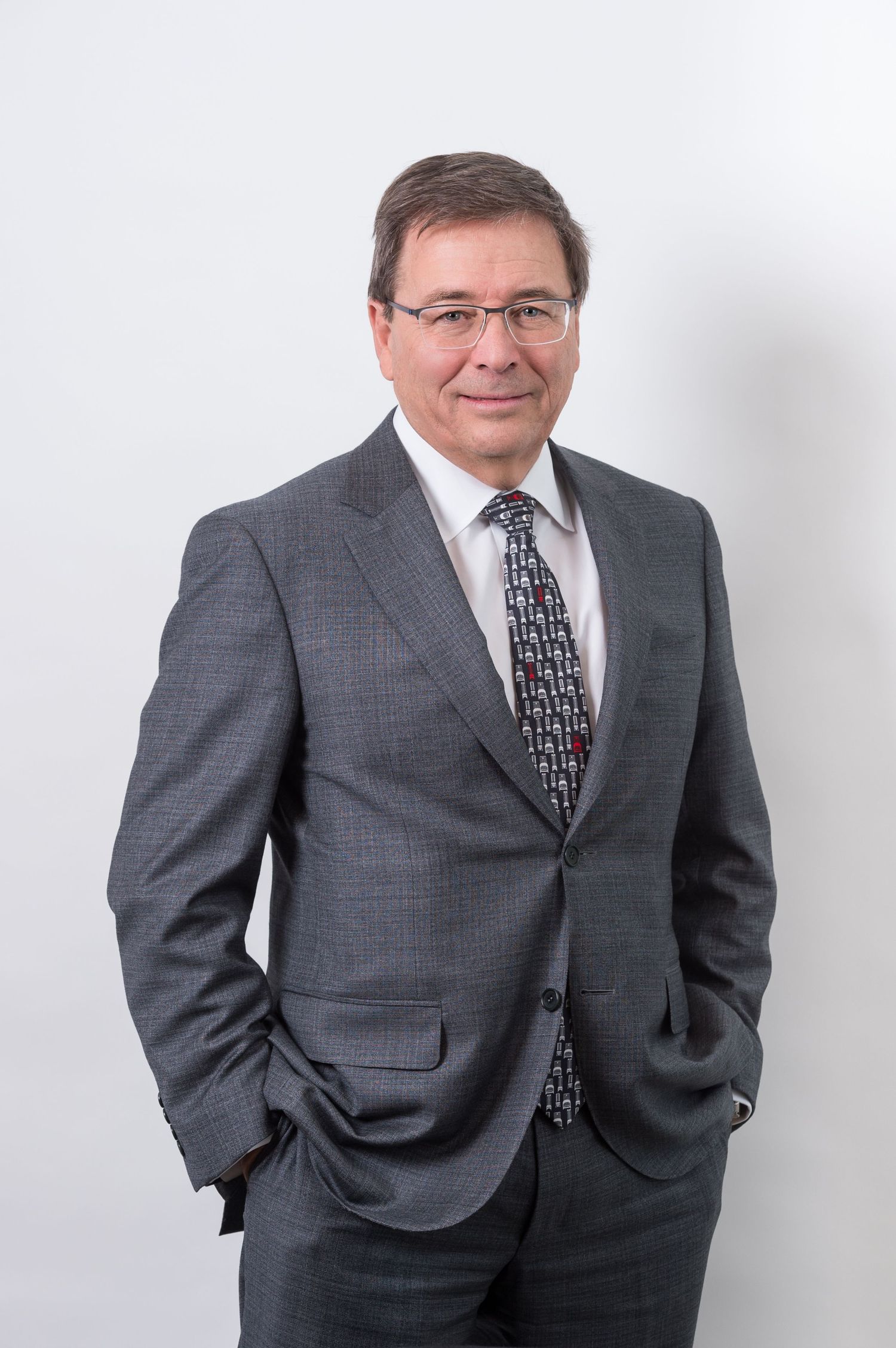Professional in grey suit and patterned tie poses against white background.