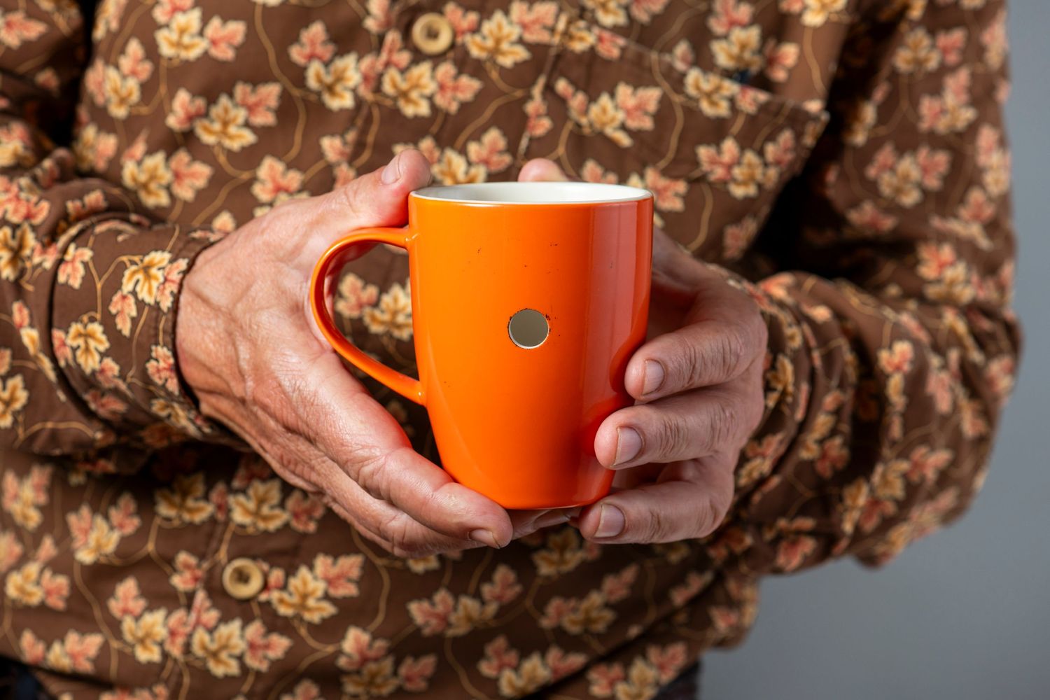 Hands holding a bright orange coffee mug against a brown floral patterned background.
