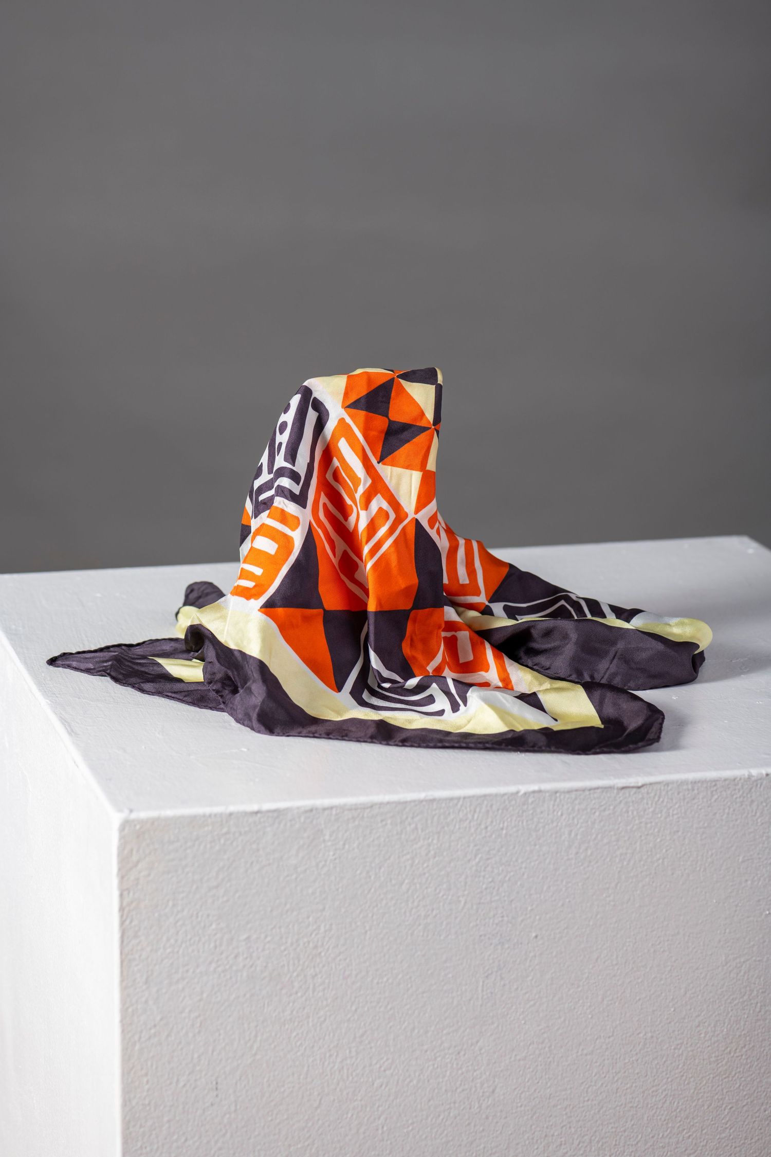 A vibrant orange and black patterned silk scarf displayed on a white pedestal against a gray background.