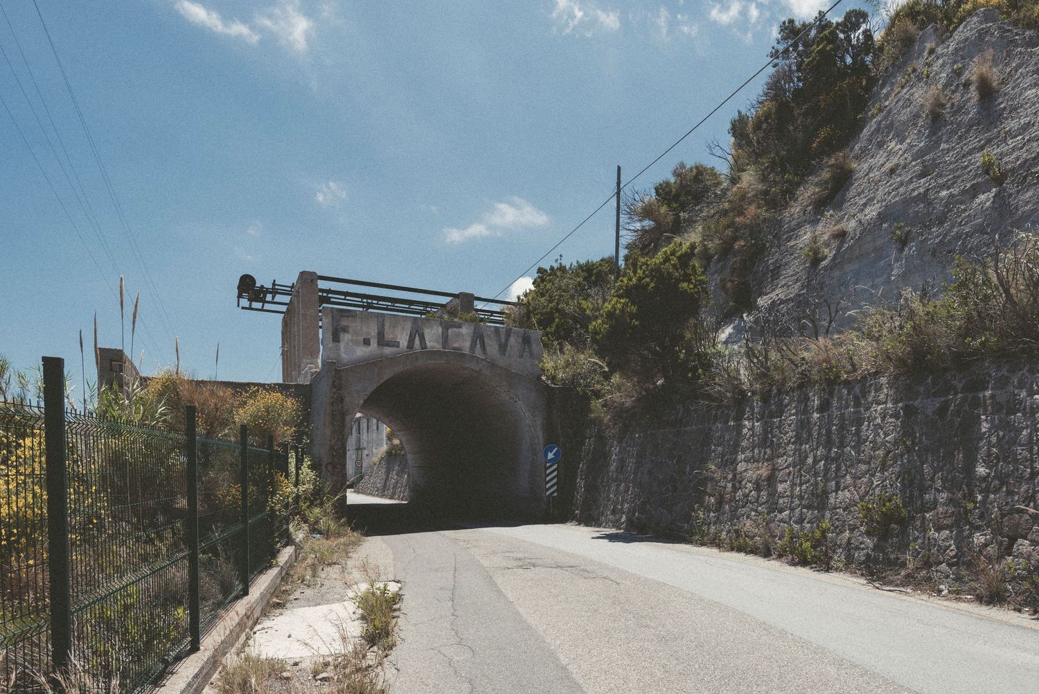Stone railway bridge tunnel entrance along a paved road with rocky cliffs and blue sky in the background.