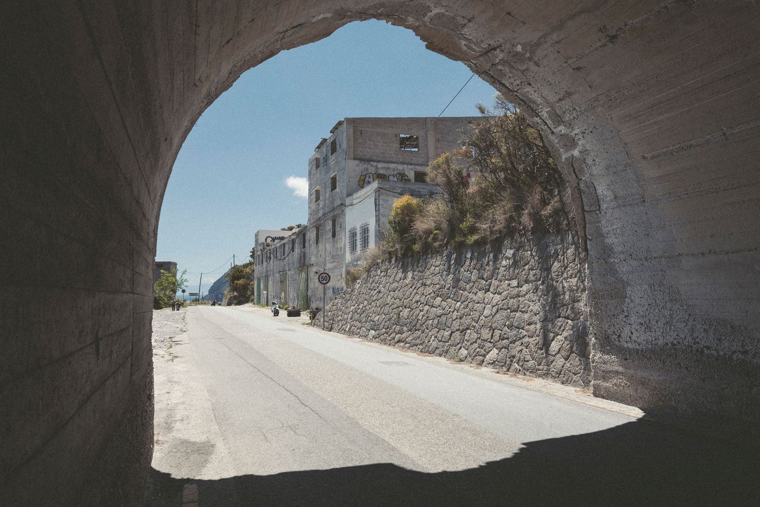 View through stone tunnel archway showing bright daylight and white buildings at the exit.