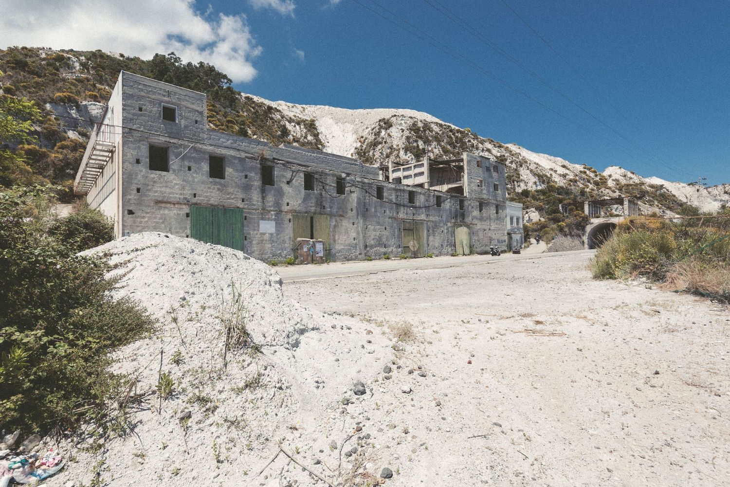 A series of abandoned stone buildings sit along a snowy mountain slope against a bright blue sky.