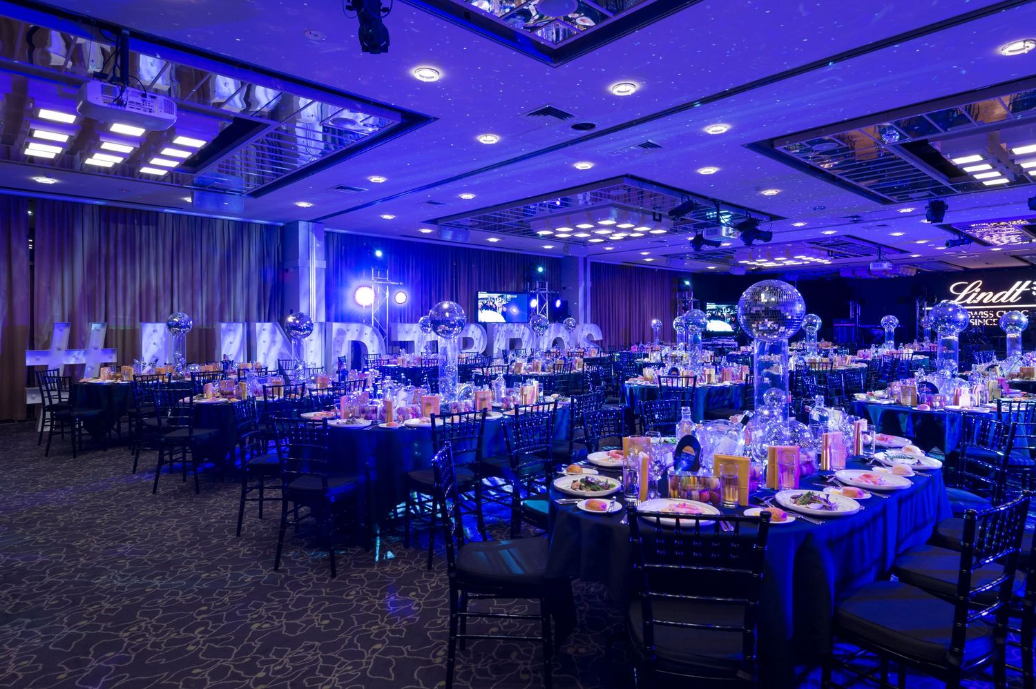 Elegant banquet hall decorated in dramatic blue lighting with round tables and formal place settings for an evening event.