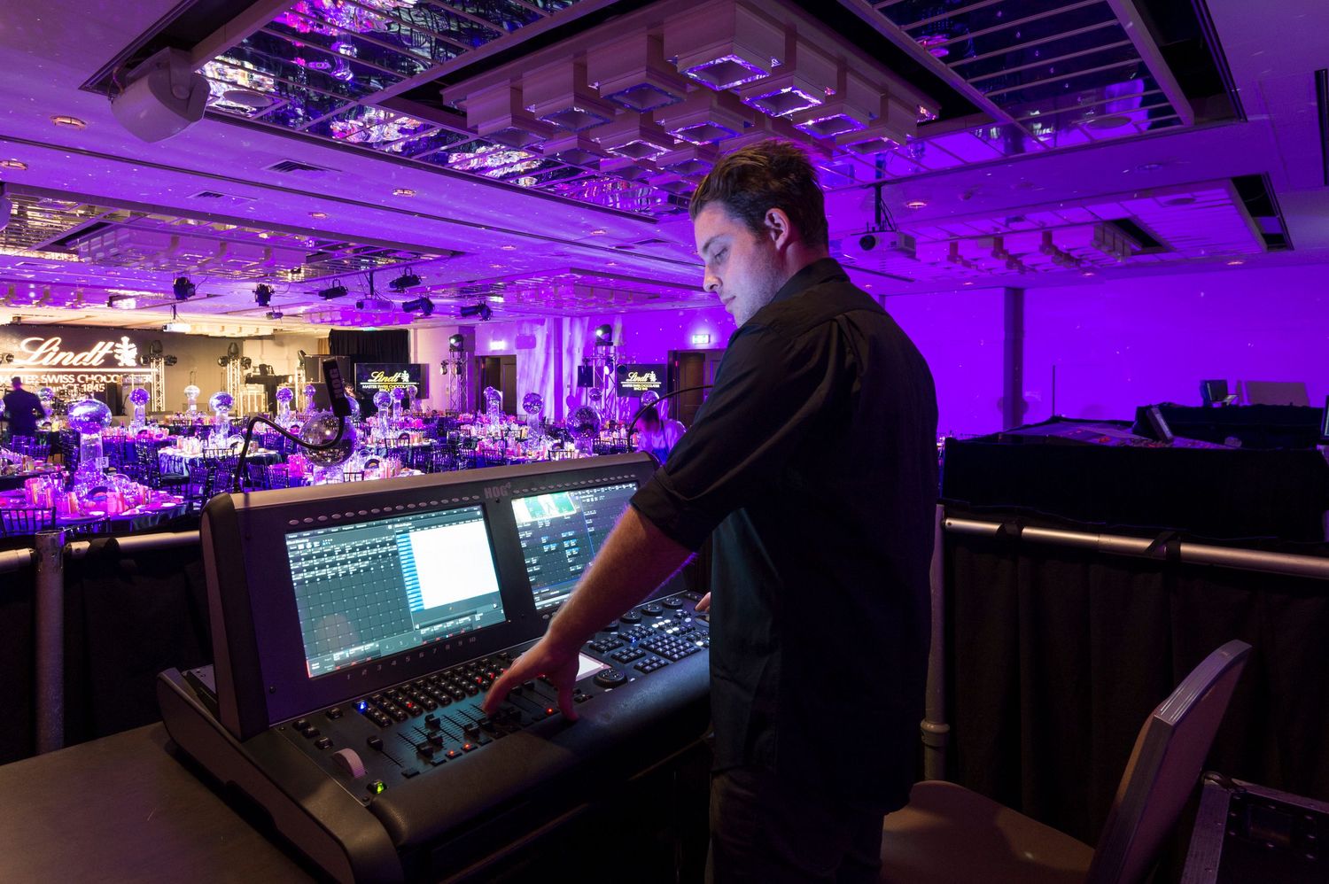 Audio engineer operates mixing console at live event venue with purple stage lighting illuminating the control booth.