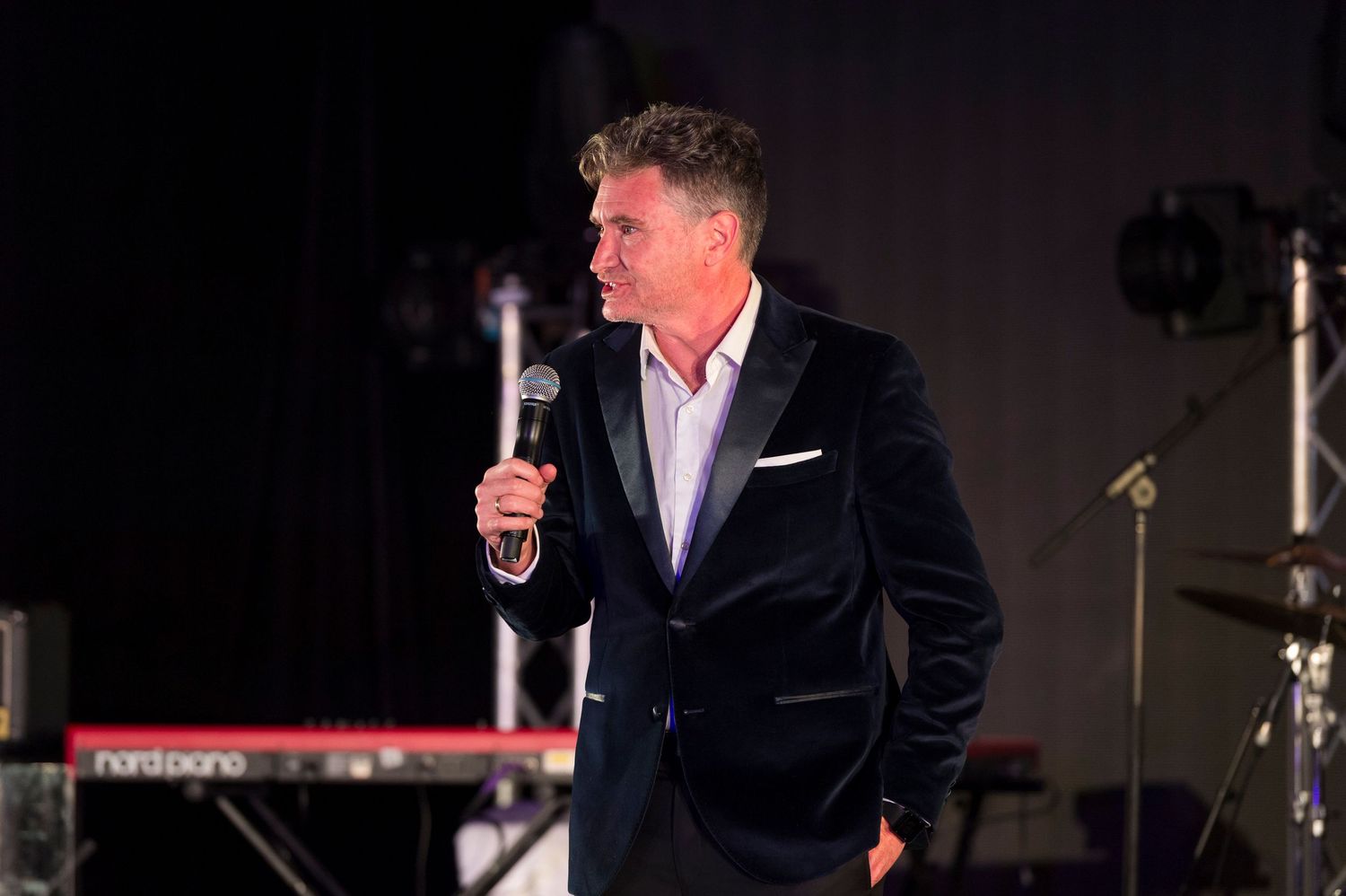 A person in a dark suit holds a glass while standing in a dimly lit room during what appears to be a formal event.