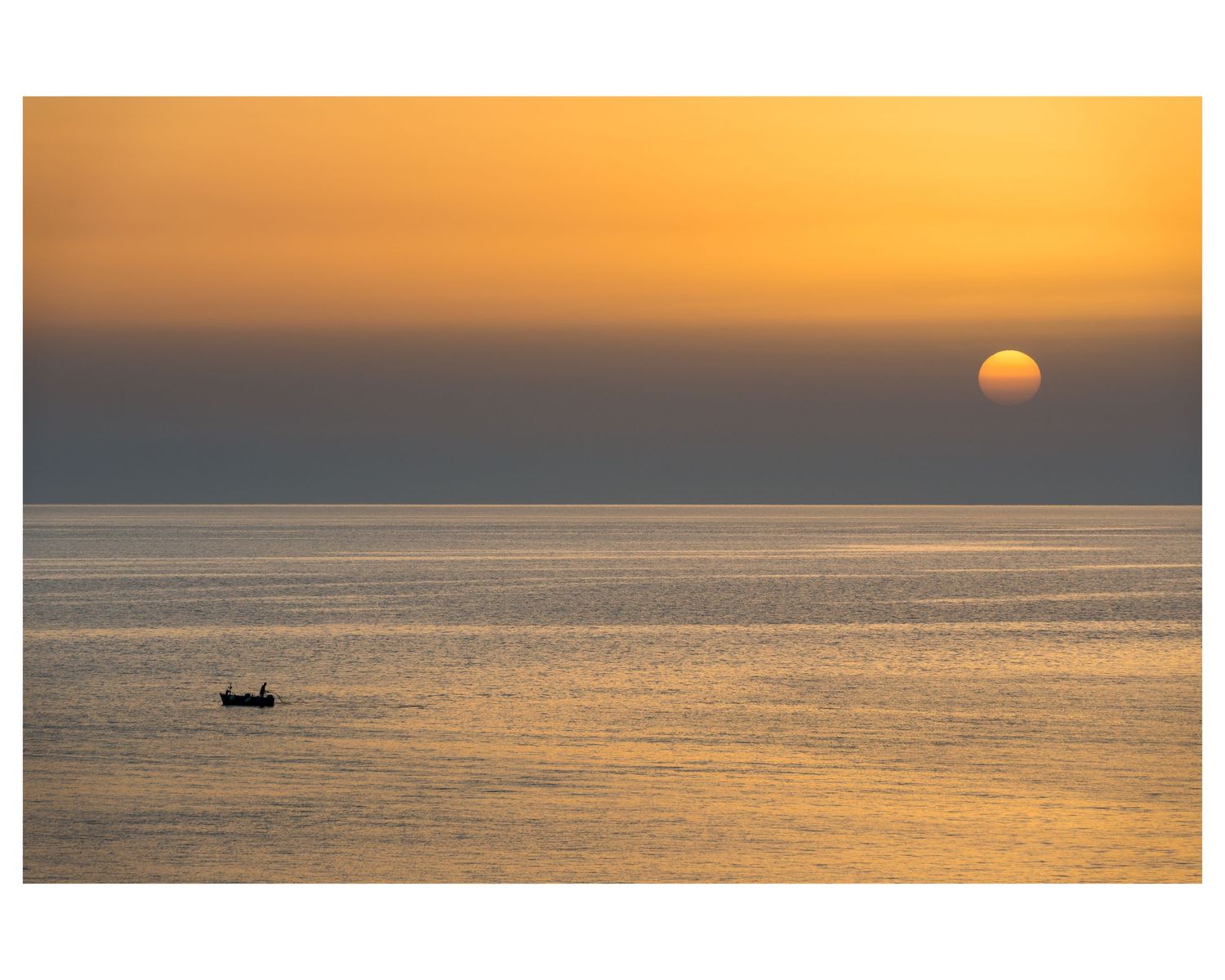 Small fishing boat silhouetted against golden sunset sky over calm ocean waters as sun dips below horizon.