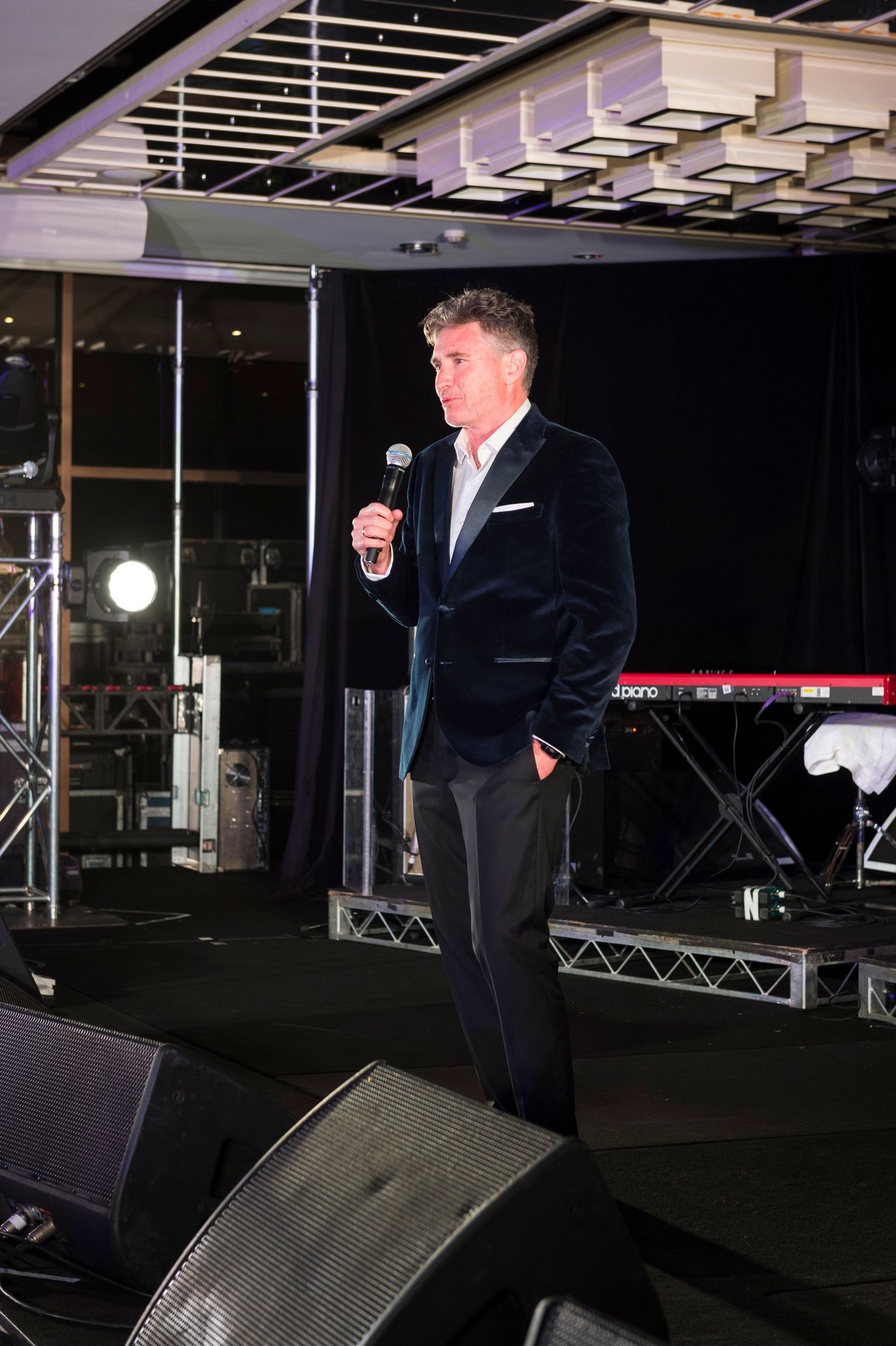 A performer in a dark suit stands on stage with a microphone at a venue with stage lighting and musical equipment.