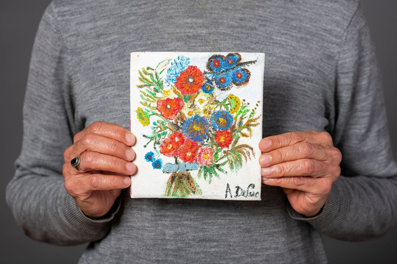 Hands holding a colorful watercolor painting of red and blue flowers with green foliage on white paper.