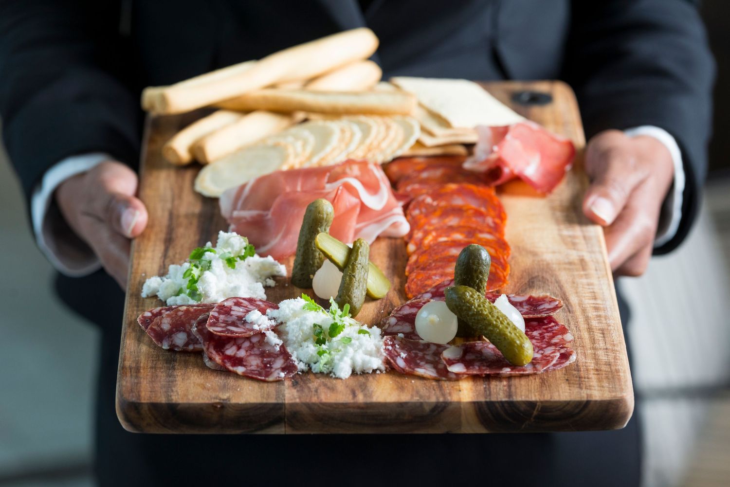 Wooden charcuterie board featuring assorted meats, cheeses, and garnishes presented on white tablecloth.