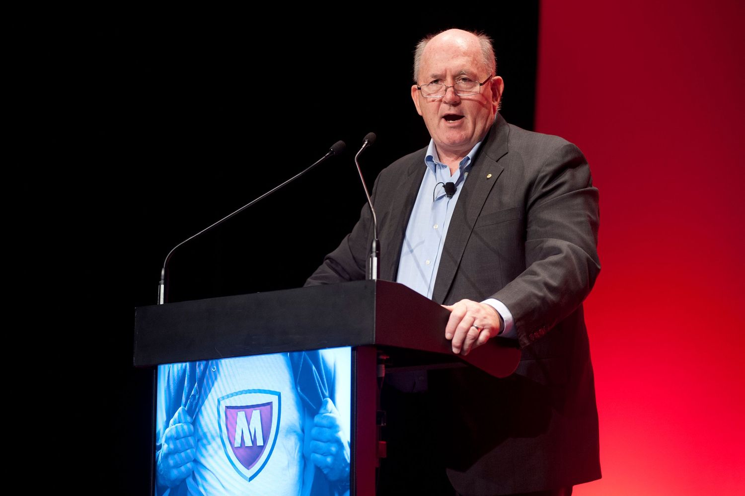 Speaker presenting at a podium on stage with red backdrop and digital displays.
