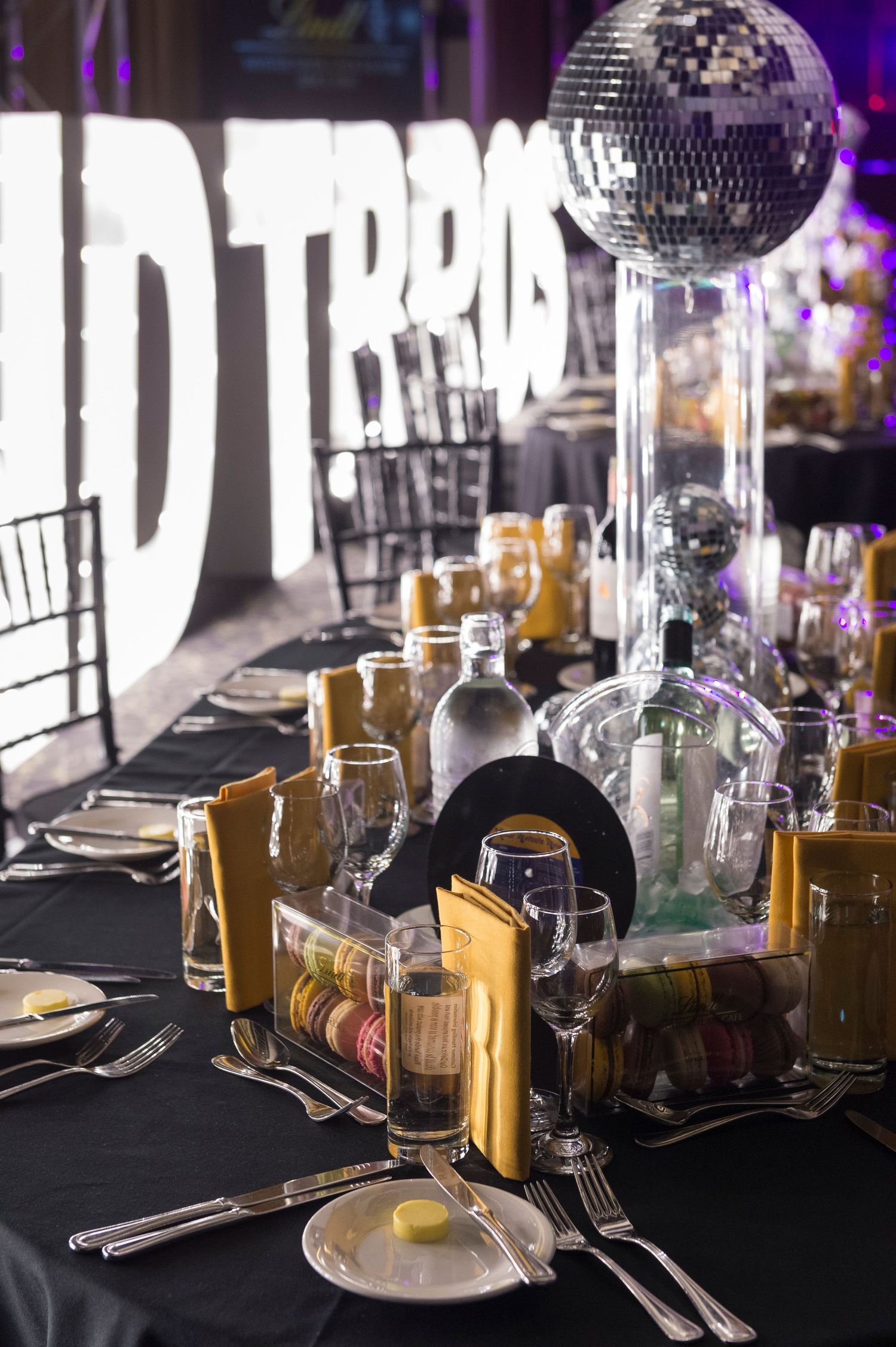 Elegant table setting with disco ball ambiance featuring black tablecloth, wine glasses and gold bottles in dim lighting.
