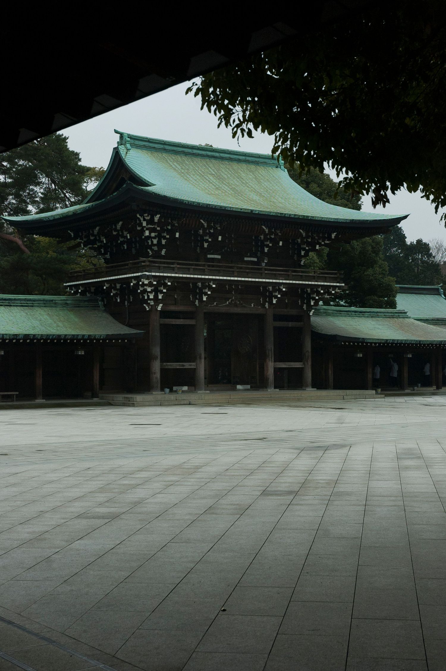 Traditional Japanese temple buildings with green tiled roofs and stone courtyard under an overcast sky.