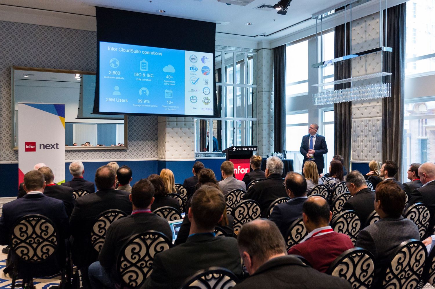 Corporate conference room filled with rows of attendees seated in ornate chairs facing presentation screens.