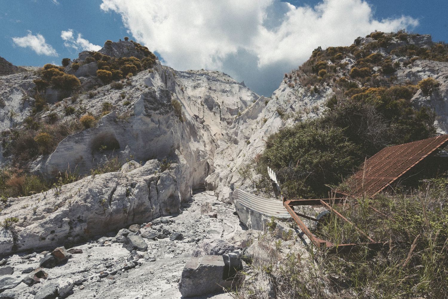 White limestone cliffs and rocky outcrops rise against blue sky with a small house nestled among trees in the mountains.