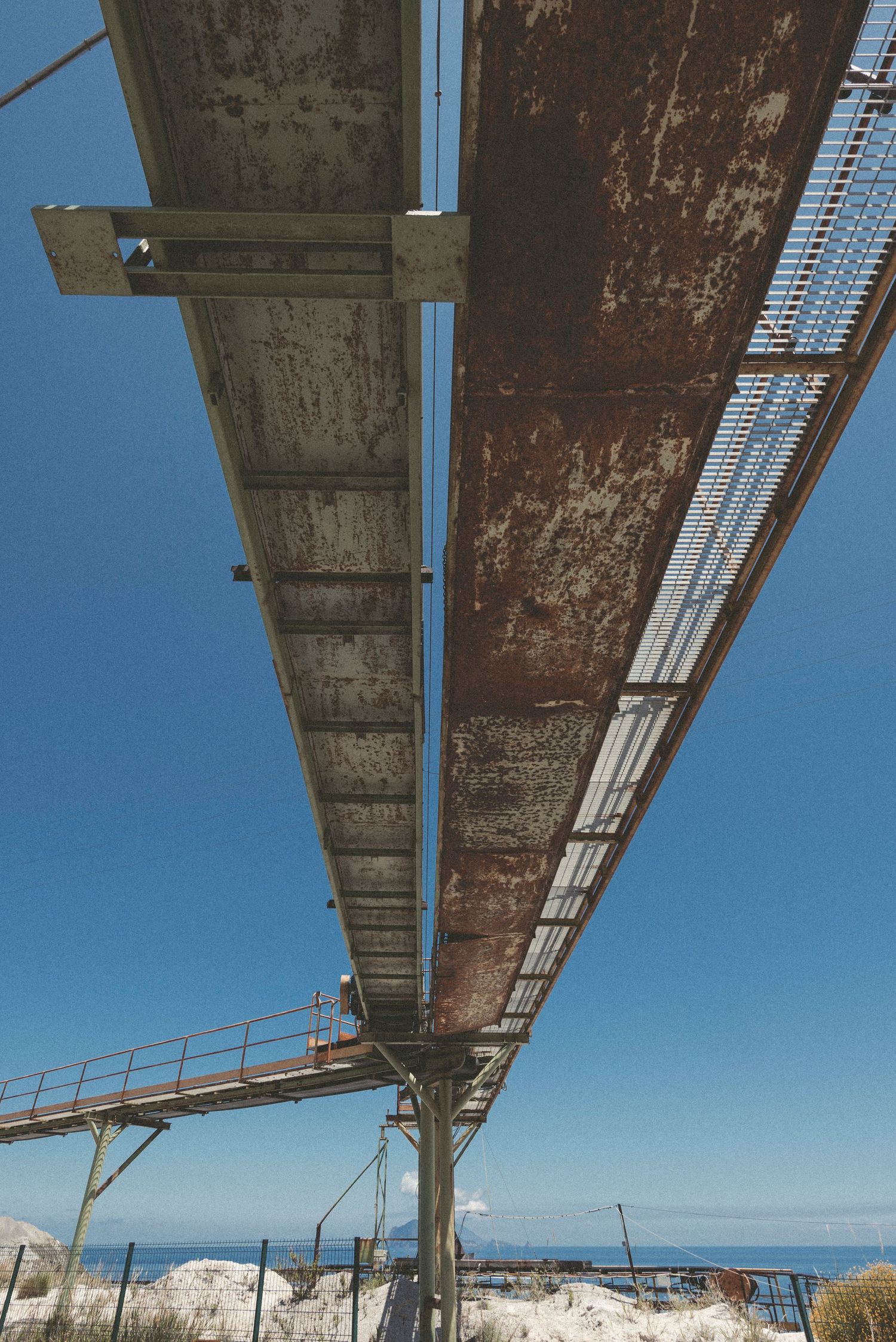 Rusty underside of an abandoned pier or bridge structure stretches across a blue sky over a coastal landscape.