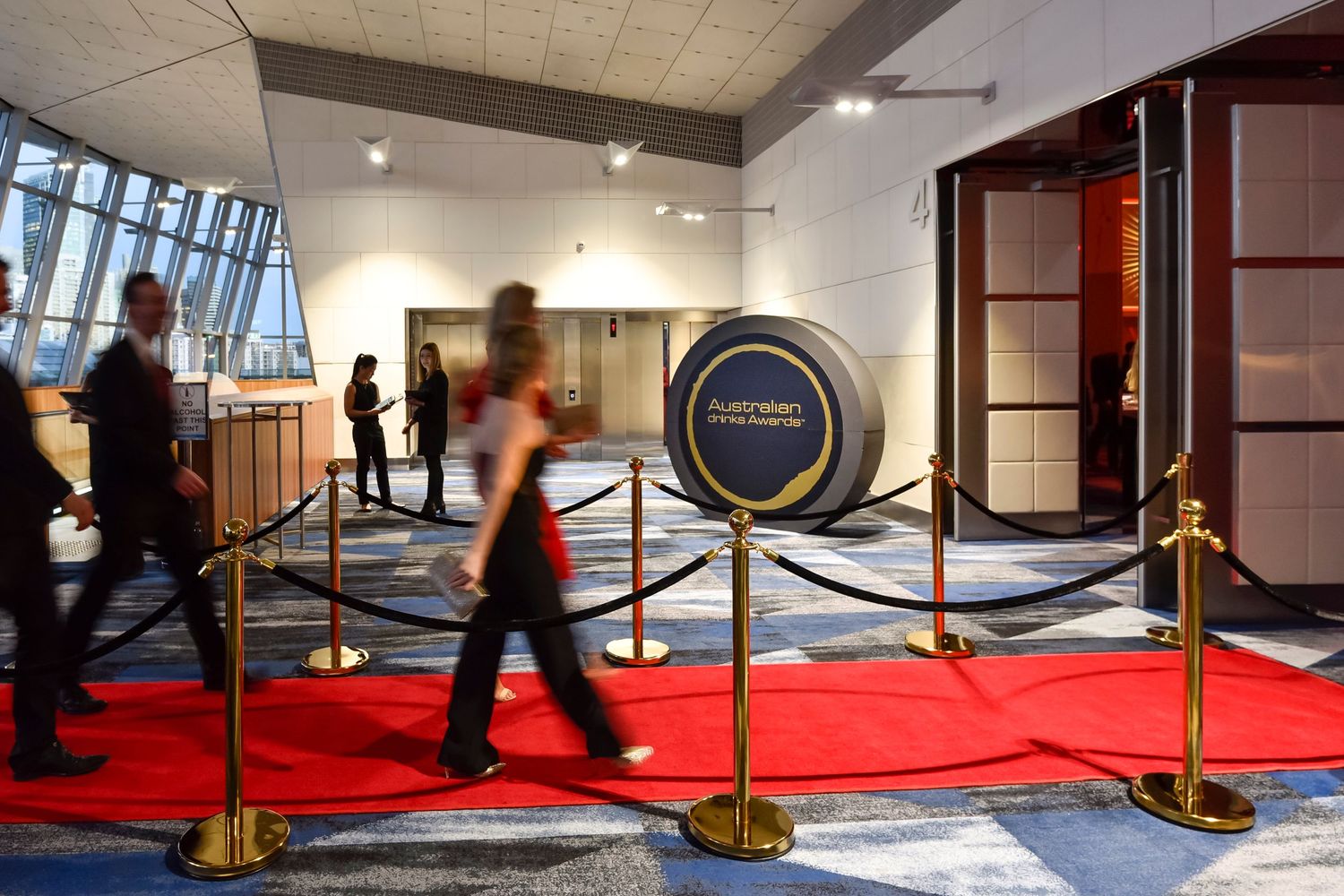 A red carpet entrance area with bollards and ropes in a modern building lobby with people walking past.