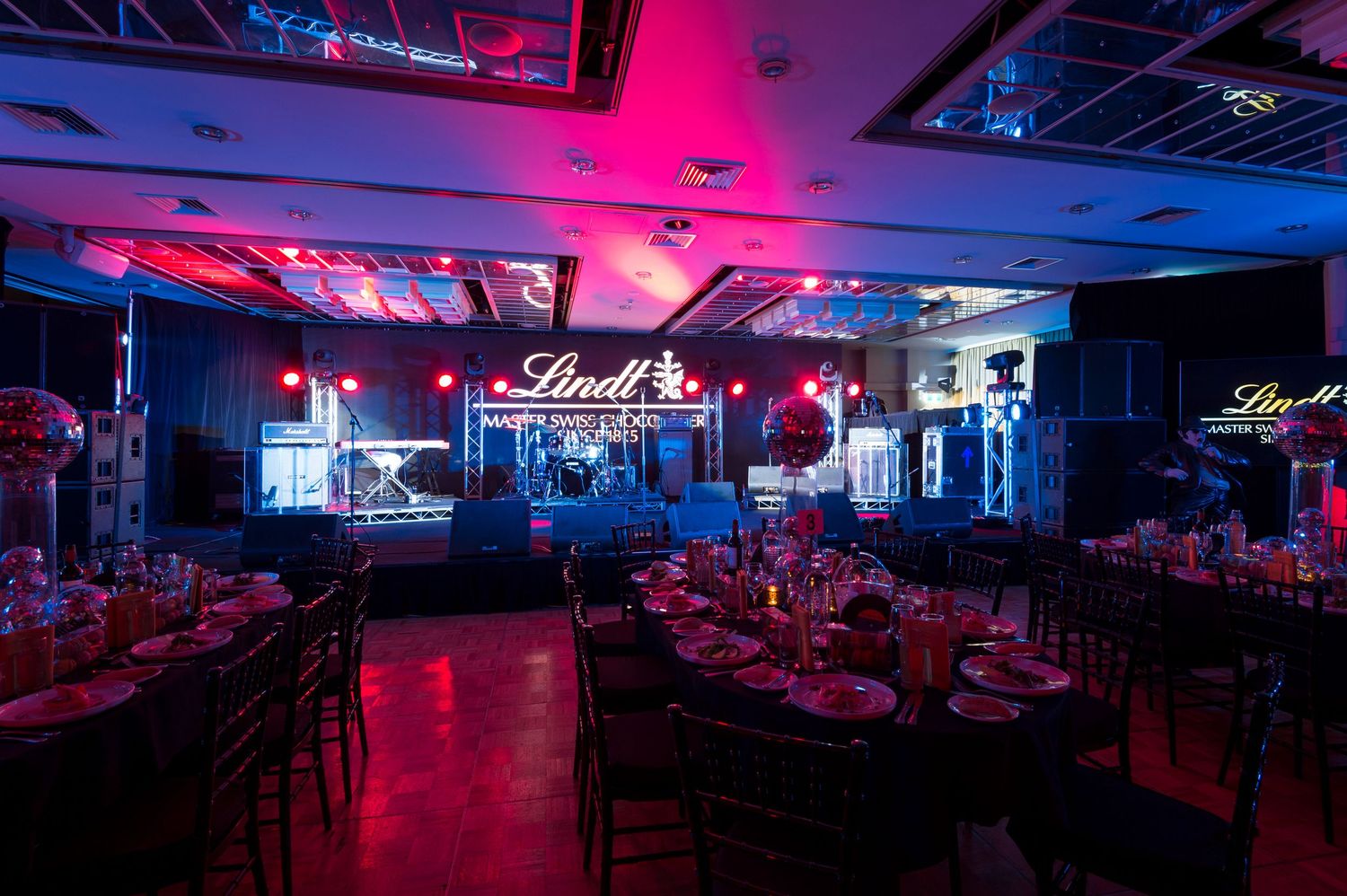 Styled room interiors set up of sponsored event with stage lighting illuminating rows of tables and chairs in a large entertainment space.