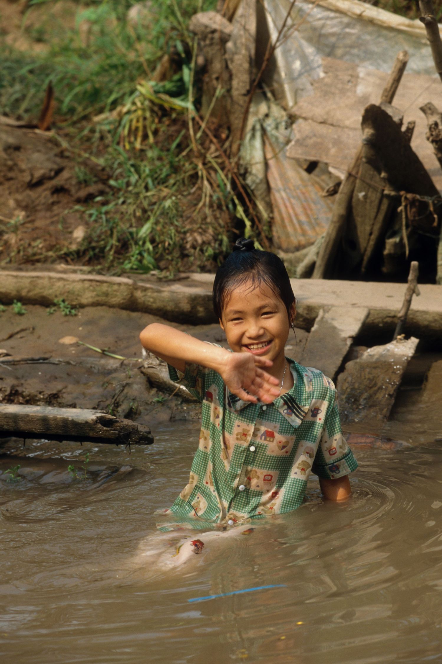 Child playing and splashing in shallow muddy river water near wooden riverbank.