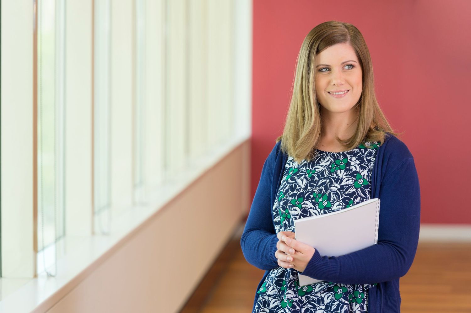 Professional wearing a floral dress and navy cardigan stands in front of red wall holding documents.