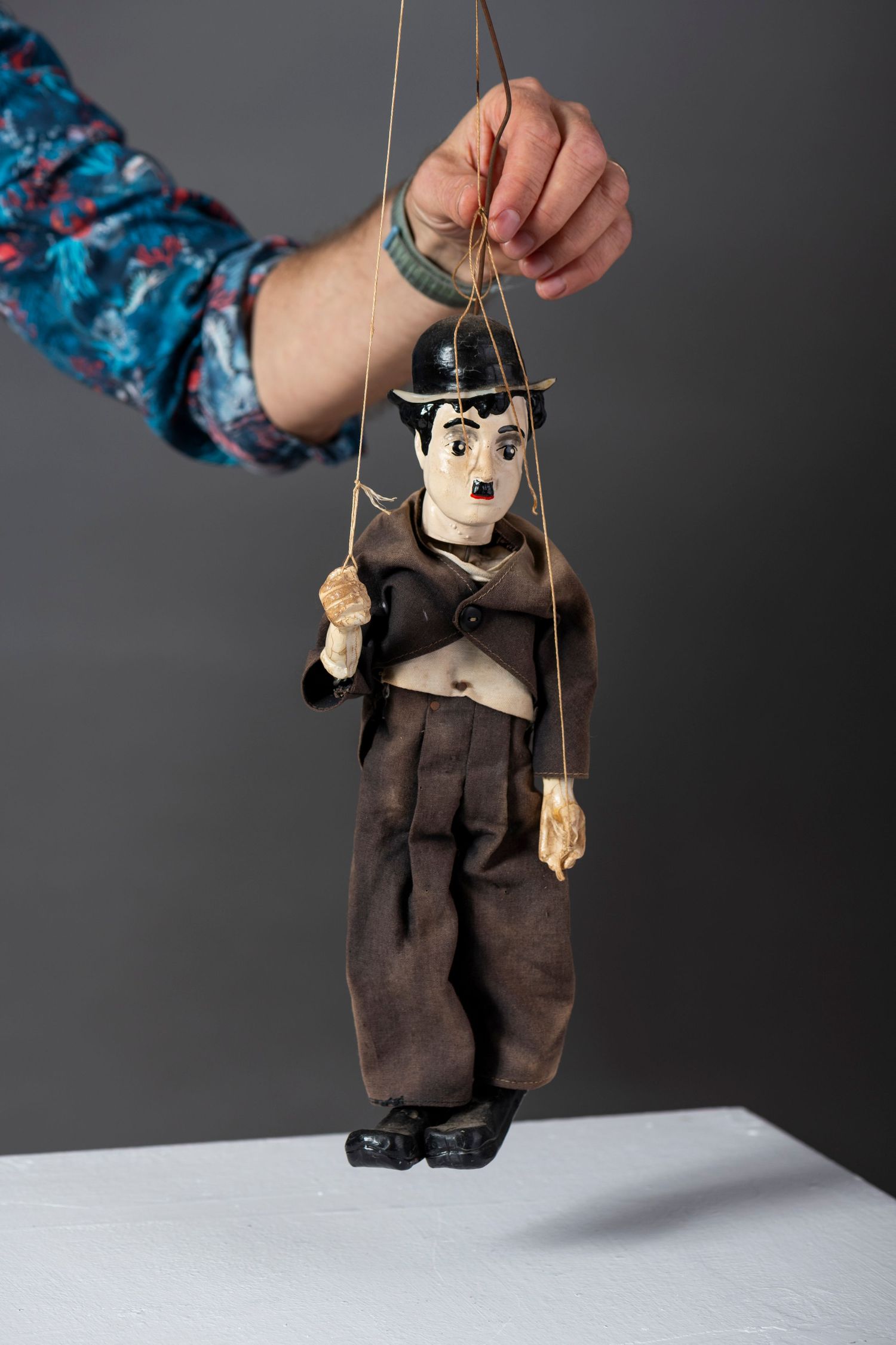 A wooden puppet marionette wearing dark clothing is suspended by strings against a gray background.
