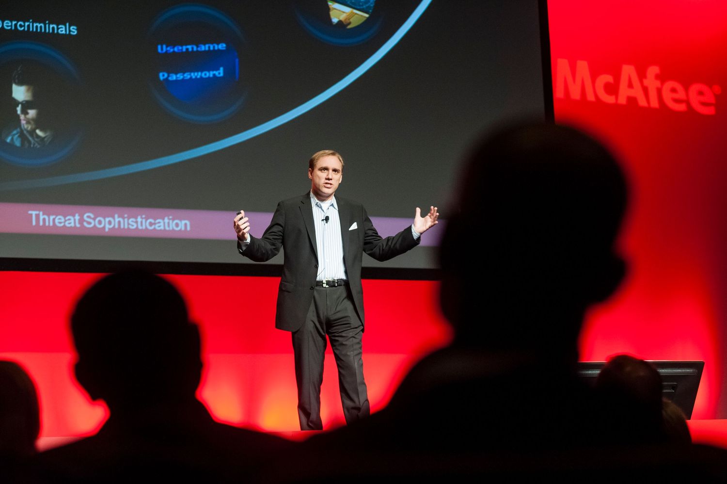 A presenter gives a speech on stage with a blue and red backdrop showing a McAfee presentation screen at Sydney ICC.