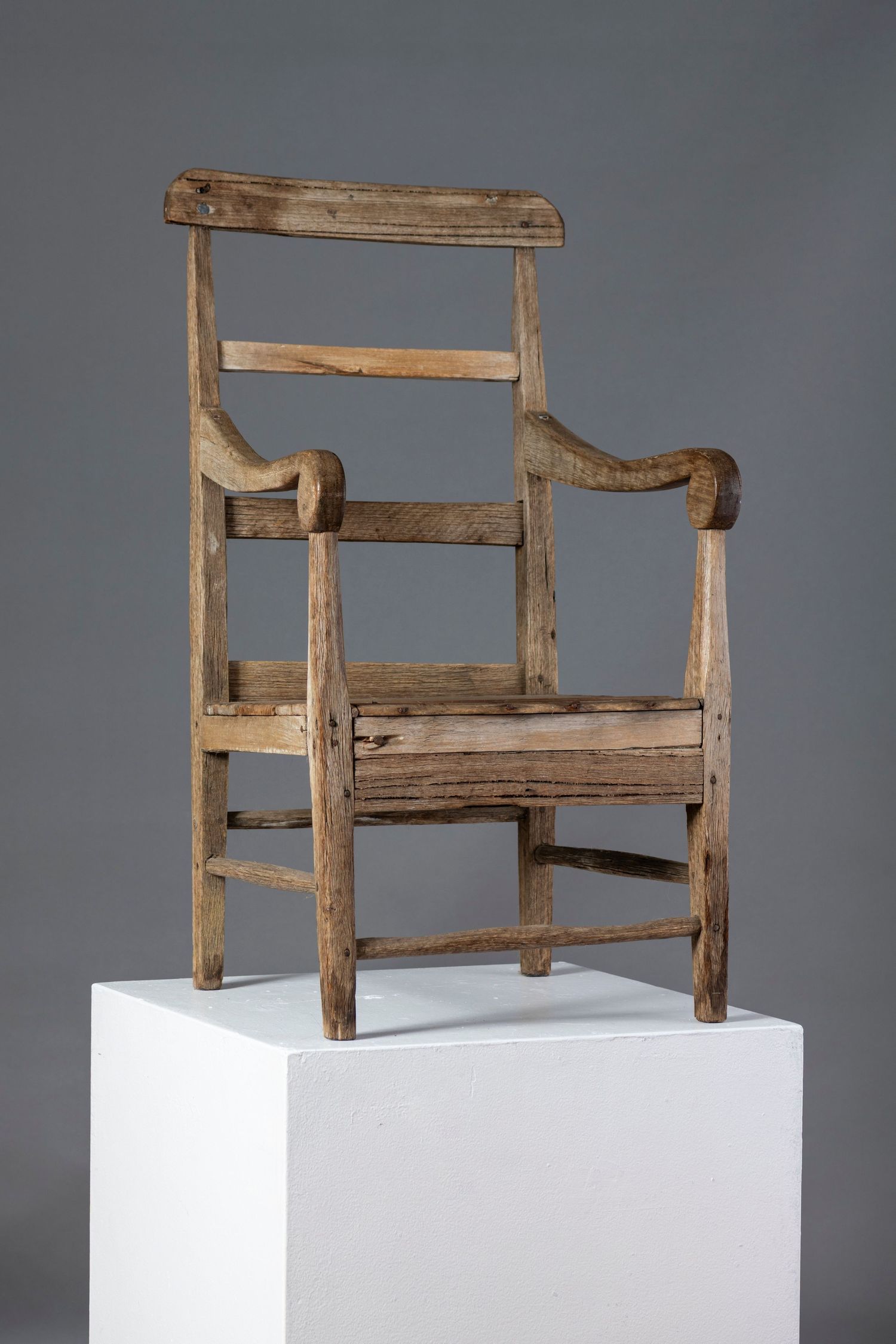 Rustic wooden armchair with slatted back and curved arms displayed on white pedestal against gray background.