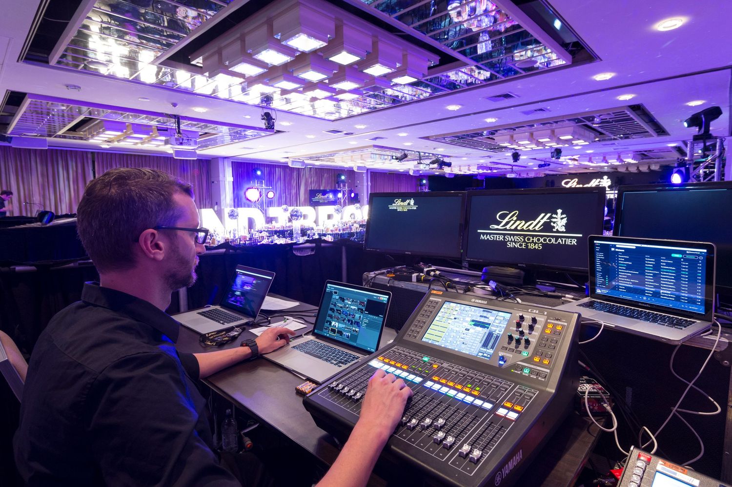 Audio technician operating a digital mixing console with purple stage lighting in a performance venue.