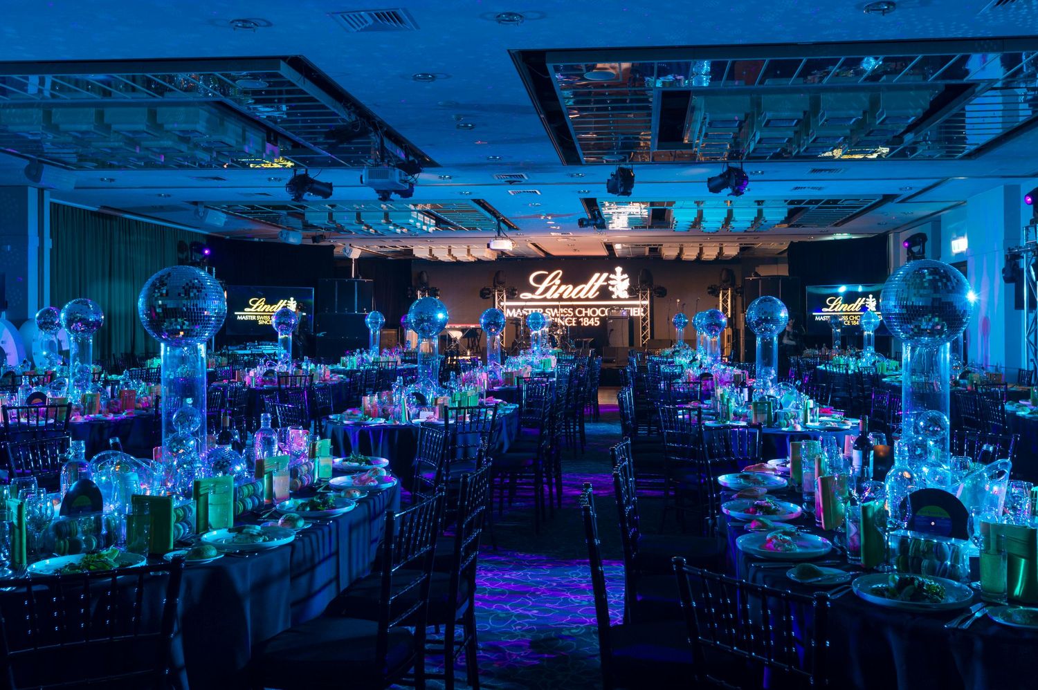 Styled venue illuminated by blue and purple neon lights for a sponsored event with stage lighting illuminating rows of table.