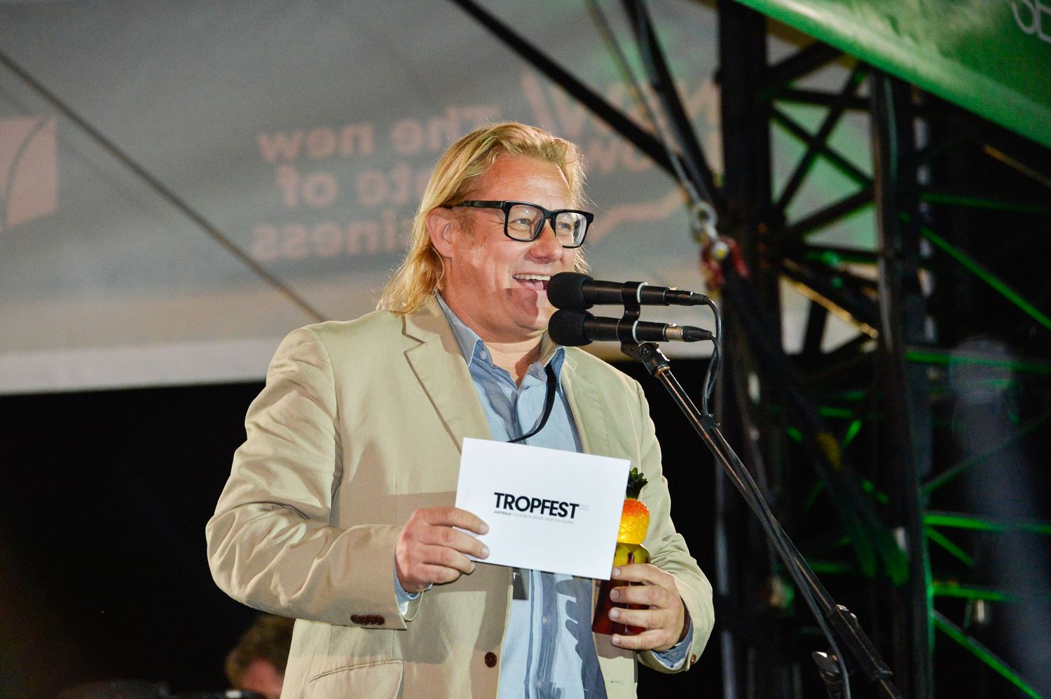 A person in a beige suit jacket speaks into a microphone while holding papers during an event.