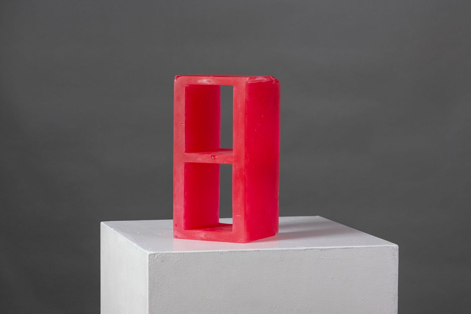 Red geometric sculpture displayed on white pedestal against dark gray background.