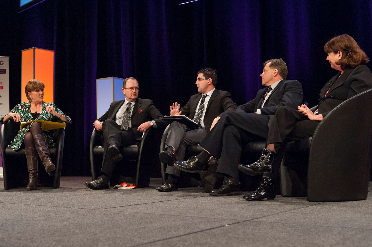 Panel discussion setup on dark stage with multiple speakers seated in chairs.