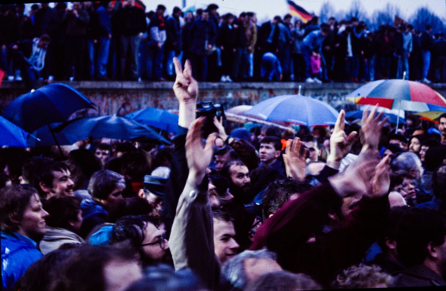 Large crowd gathered outdoors at dusk with raised hands and umbrellas visible against blue lighting.