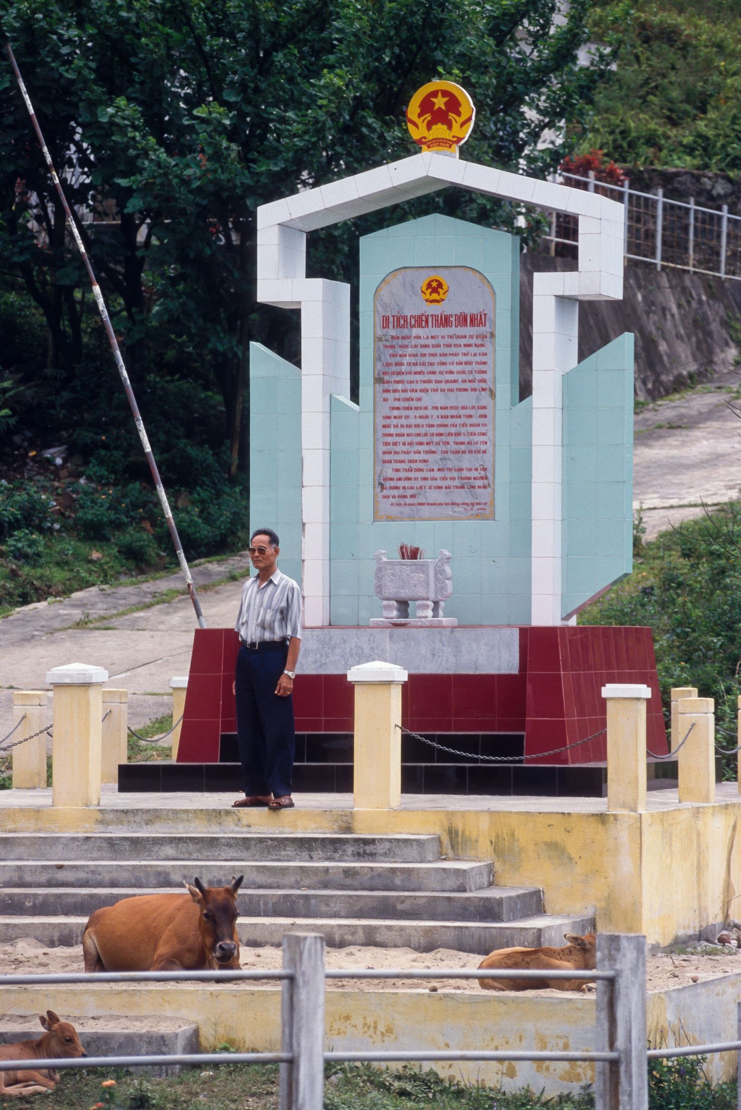 Simple concrete monument with Vietnamese text and national emblem stands along rural path.