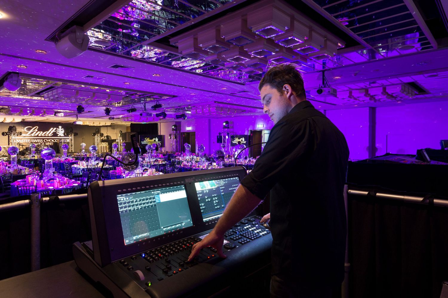 Purple-lit control room with mixing consoles and monitors showing audio production equipment in a professional studio setting.