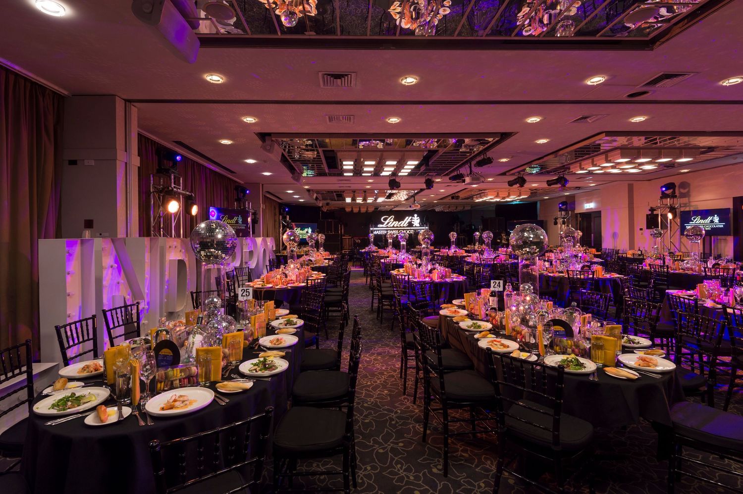An elegant banquet hall with round tables set for a formal event, illuminated by purple and pink ambient lighting.