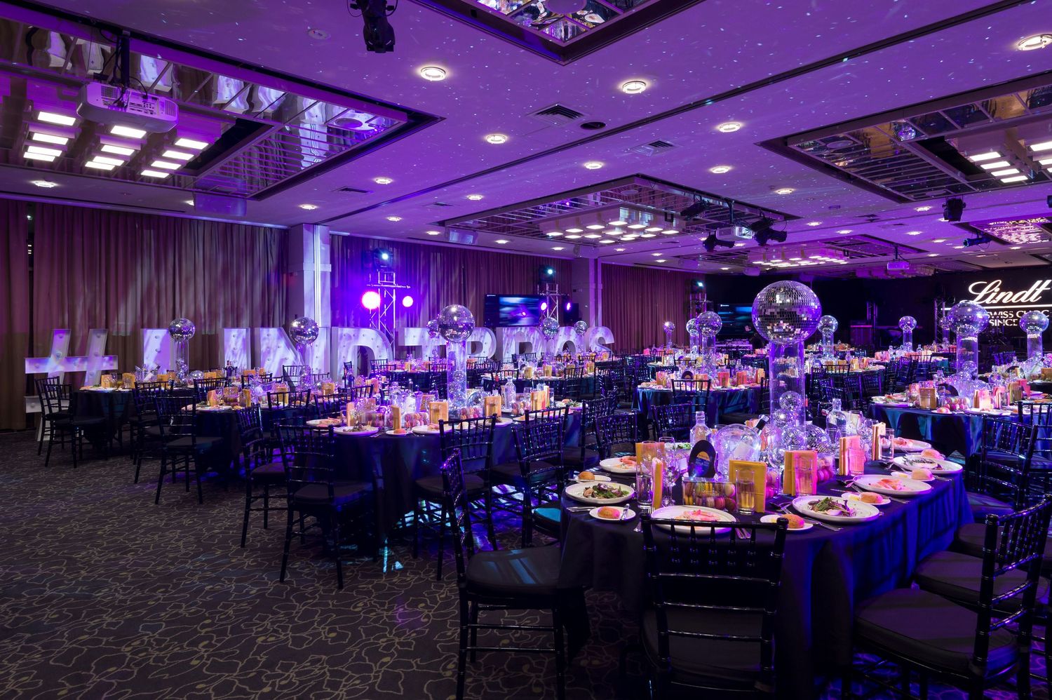 Large ballroom with purple uplighting and elegant table settings arranged for a formal event or reception.