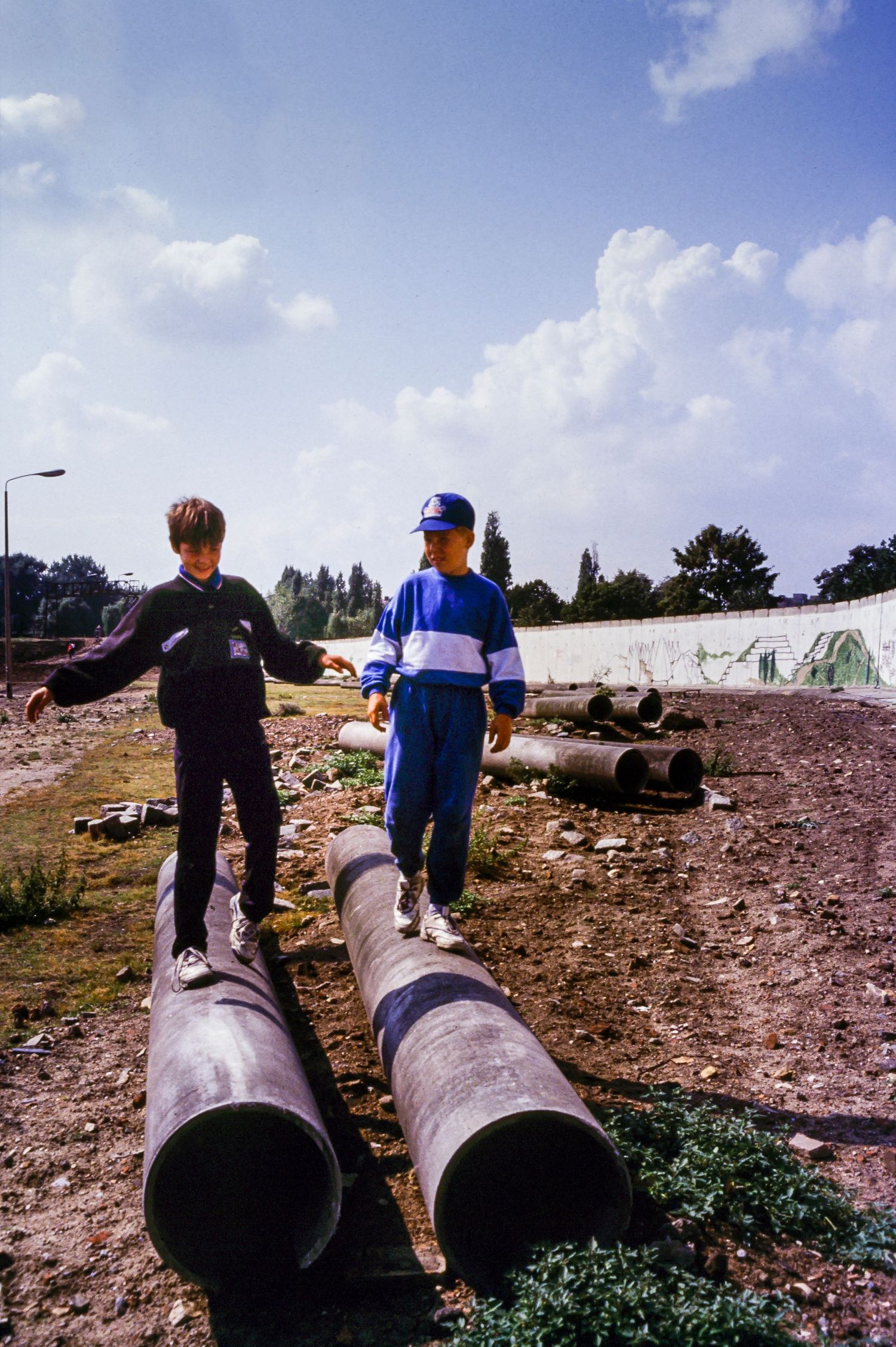 People walking along large concrete pipes near the fallen Berlin Wall.